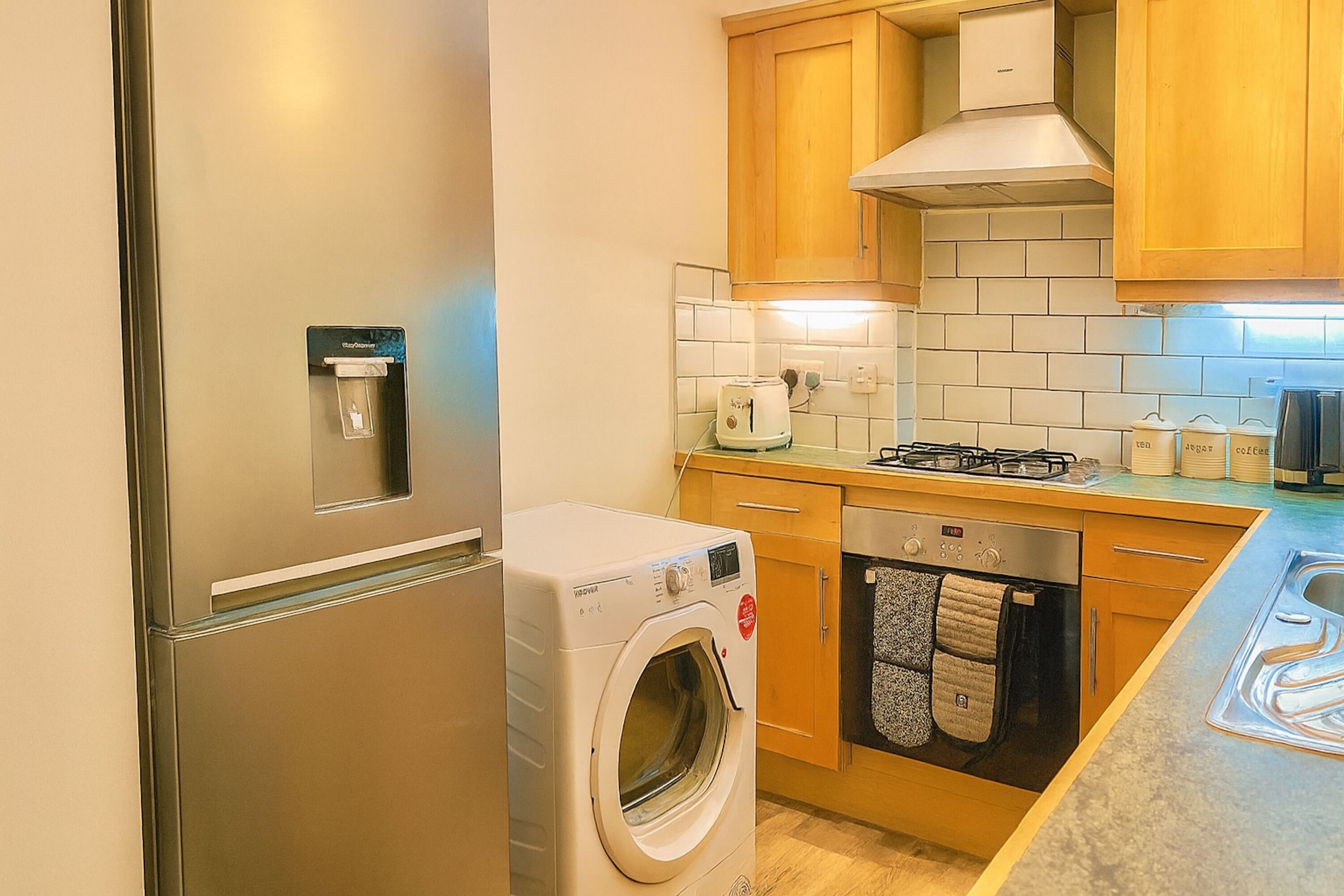 A compact kitchen with a built-in washer and dryer, wooden cabinets, a stainless steel refrigerator, and a tiled backsplash.