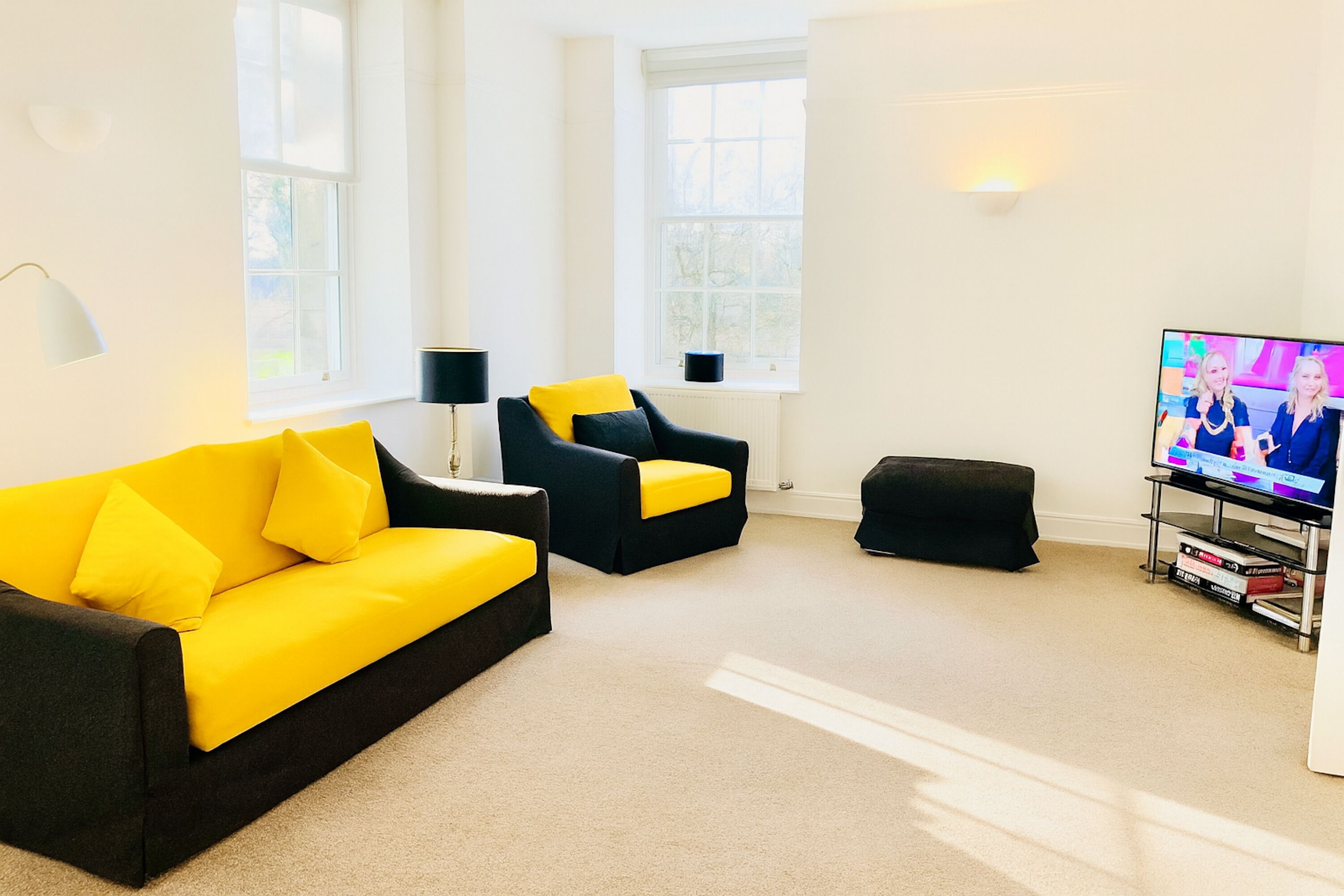 Una sala de estar moderna con un sofá amarillo, un sillón negro y un reposapiés negro. Un televisor está a la derecha.
