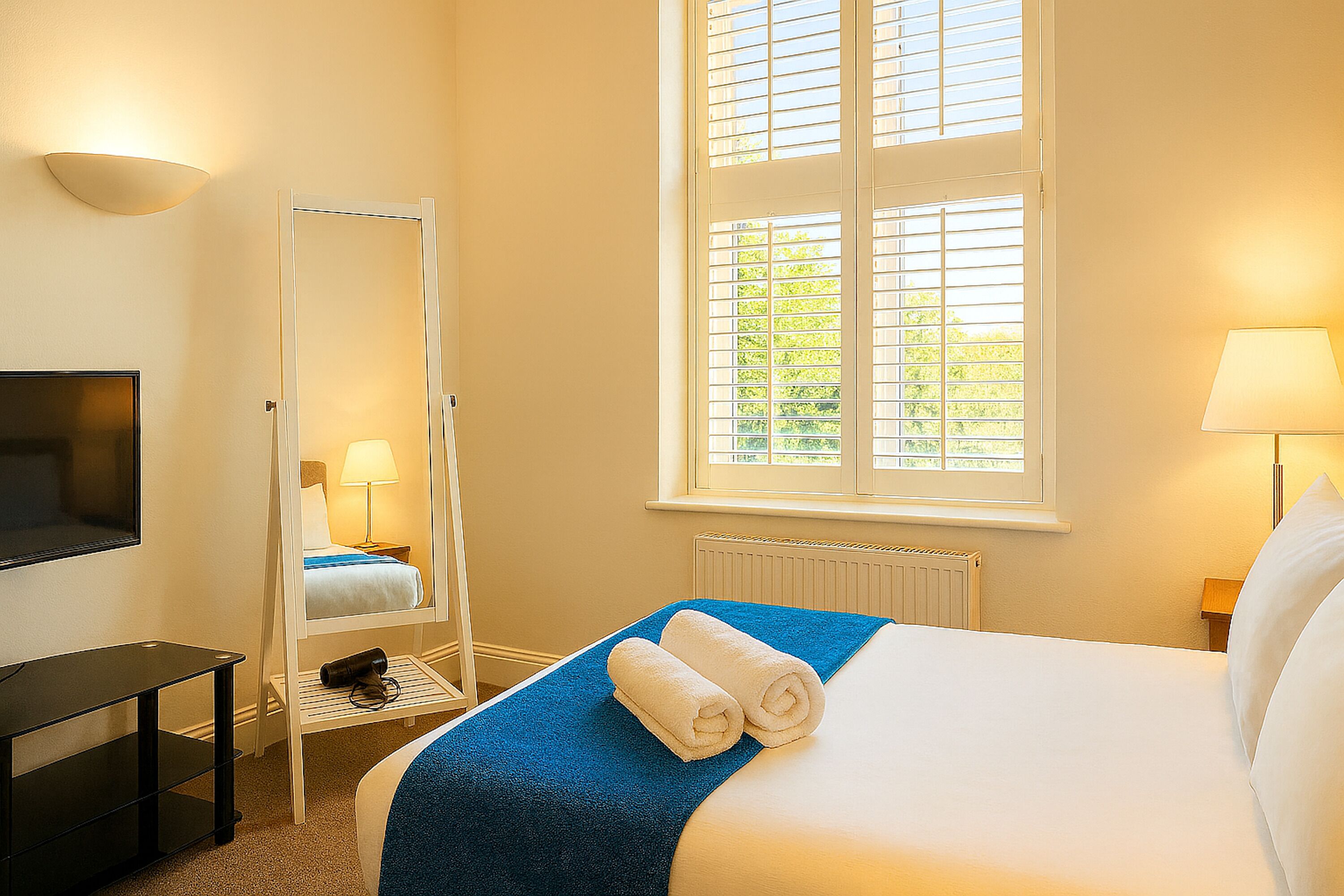 A hotel room with a bed, a TV, a mirror, a window with blinds, and a lamp.