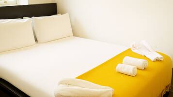 A neatly made bed with white linens, a yellow throw blanket, and rolled towels on the bed.