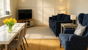 Uma sala de estar moderna com uma televisão de tela plana, um sofá azul escuro, uma mesinha com um abajur e um vaso de flores amarelas sobre uma mesa branca.