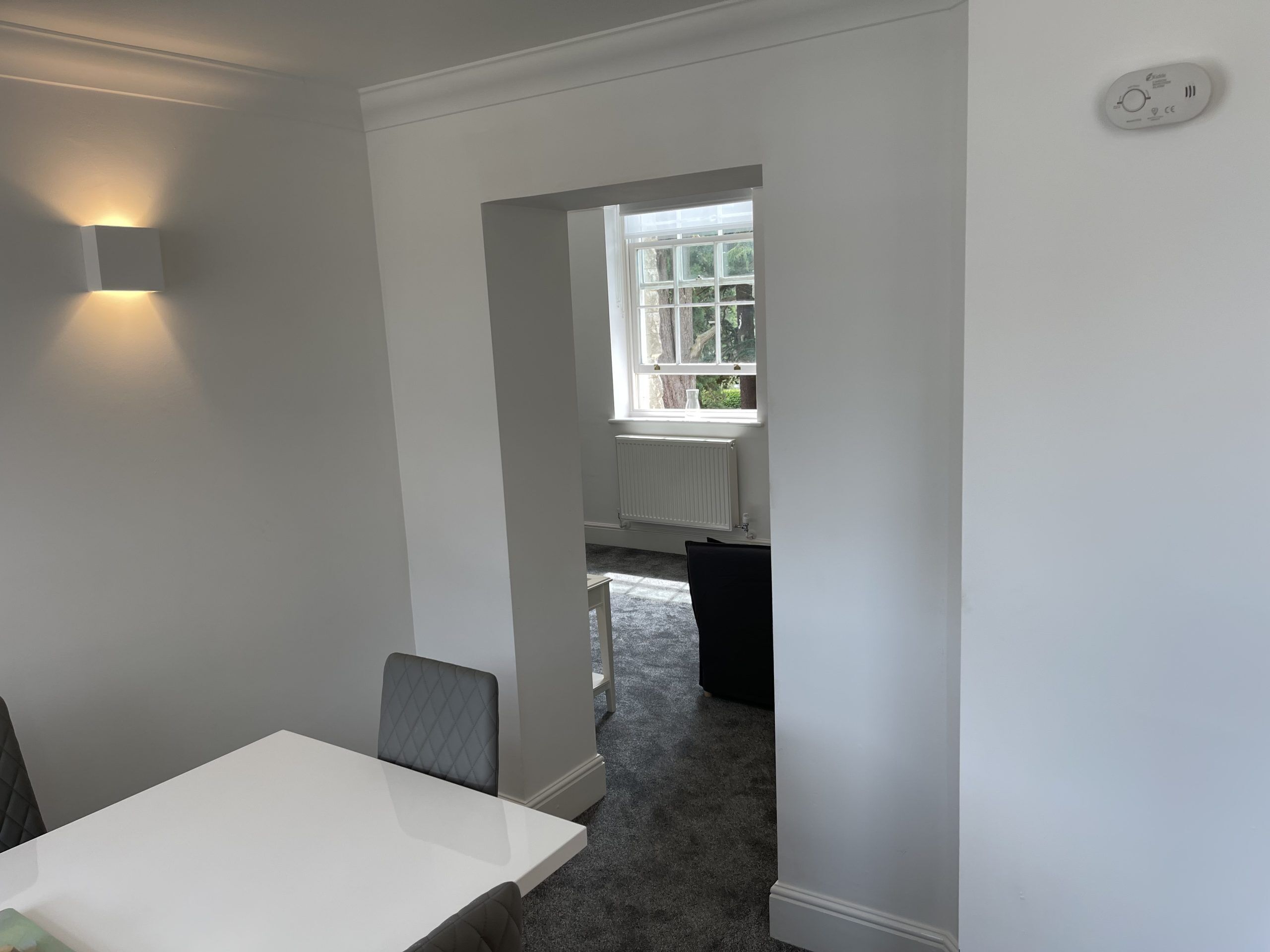 Une pièce avec une table et des chaises blanches, un radiateur et une fenêtre donnant sur un paysage verdoyant.