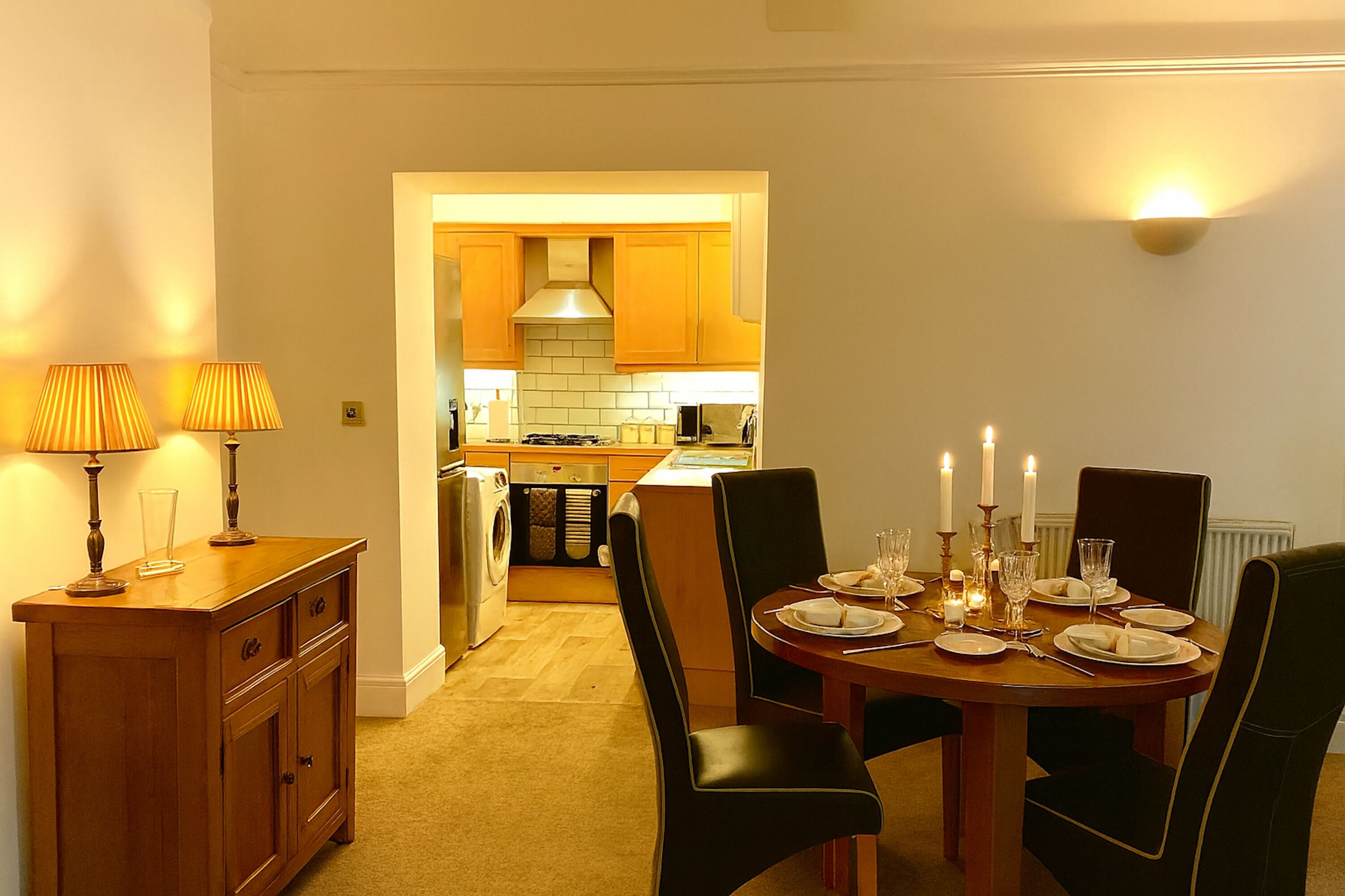 Un comedor con una mesa de madera dispuesta para cuatro personas, rodeada de sillas negras. A la izquierda, un mueble de madera con una lámpara. Se ve la cocina al fondo, con armarios de madera y una estufa.