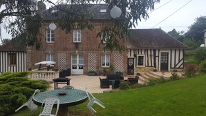 Une maison traditionnelle à colombages, dotée d’une façade en briques, d’un porche avec un espace salon extérieur et d’un jardin agrémenté d’une table ronde et de chaises.
