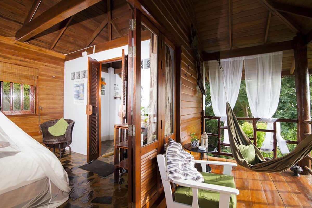 Eine Holzhütte mit einem Bett, einem kleinen Tisch mit Lampe, einer Hängematte und Blick ins Grüne.