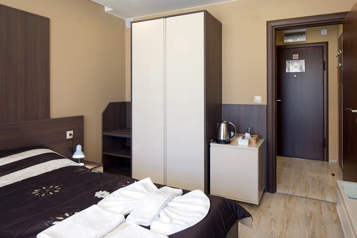 A hotel room with a bed, a wardrobe, a small cabinet with a kettle, and a door with a no-smoking sign.