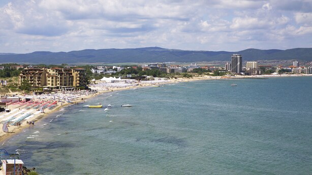 Paisatge urbà costaner amb platja, edificis i muntanyes al fons.