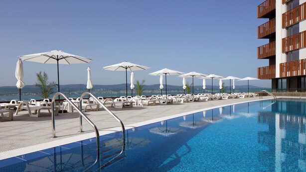 A pool area with lounge chairs and umbrellas, a clear sky, and a view of distant mountains. - Paradiso Apartcomplex (Nessebar)