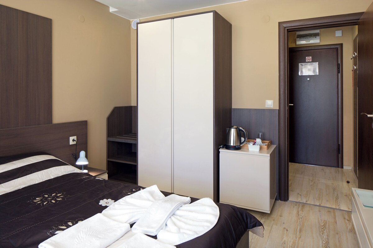 Une chambre d’hôtel avec un lit, une armoire, un bureau avec une bouilloire et une porte avec une pancarte.