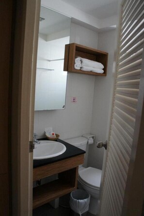 Baño pequeño con lavabo blanco, inodoro y espejo sobre el lavabo. Hay un estante de madera con toallas encima del espejo.