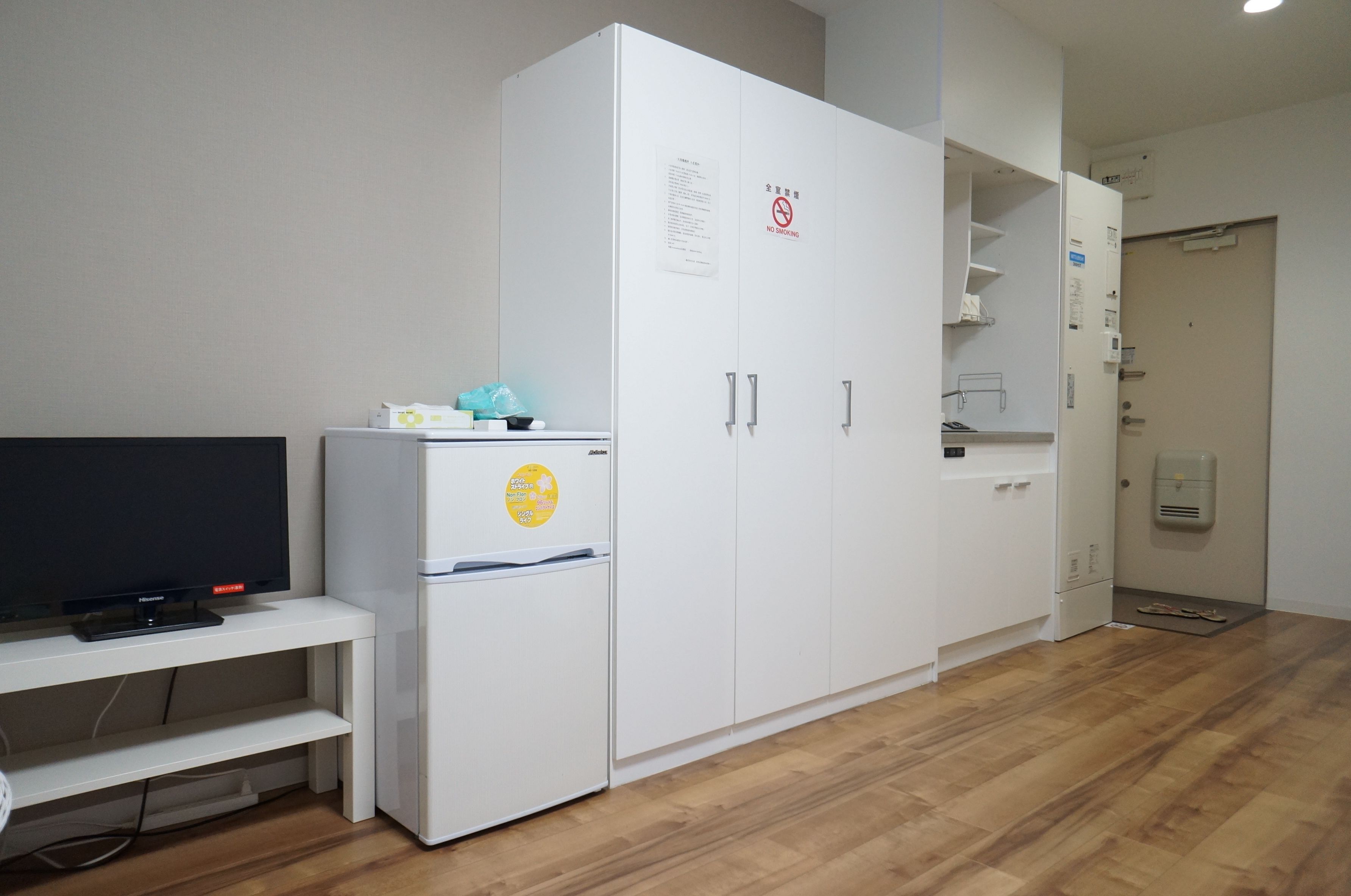 A room with a TV on a stand, a white wardrobe, a small fridge, and a wooden floor.
