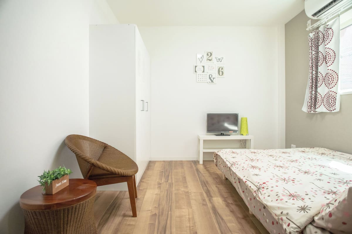 Un dormitorio con una cama, una silla, una mesita con una planta, un televisor sobre un mueble bajo y una pared con números decorativos.