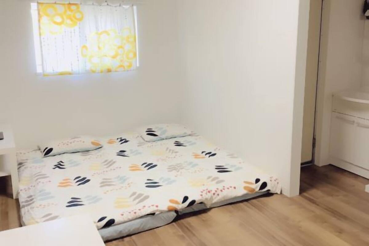 Una habitación pequeña y luminosa con una cama, una ventana con cortinas y una pared blanca.