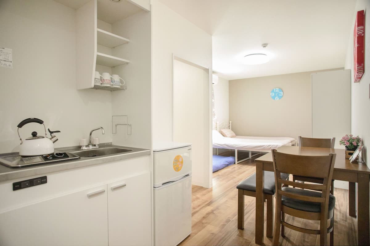 Una cocina compacta con hervidor, pileta y heladera pequeña. Hay una zona de comedor con mesa y sillas, y una cama con un reloj de pared azul.