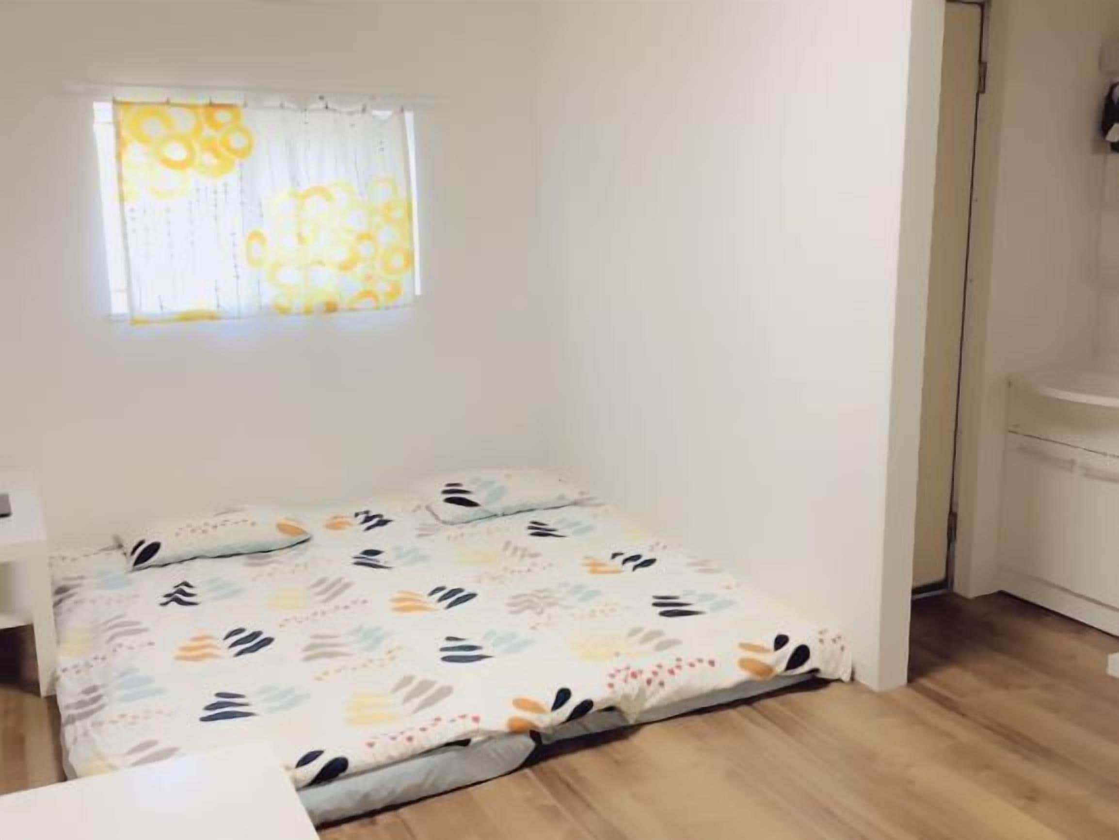 A small, single-bed room with a window, a bedside table, and a wooden floor.