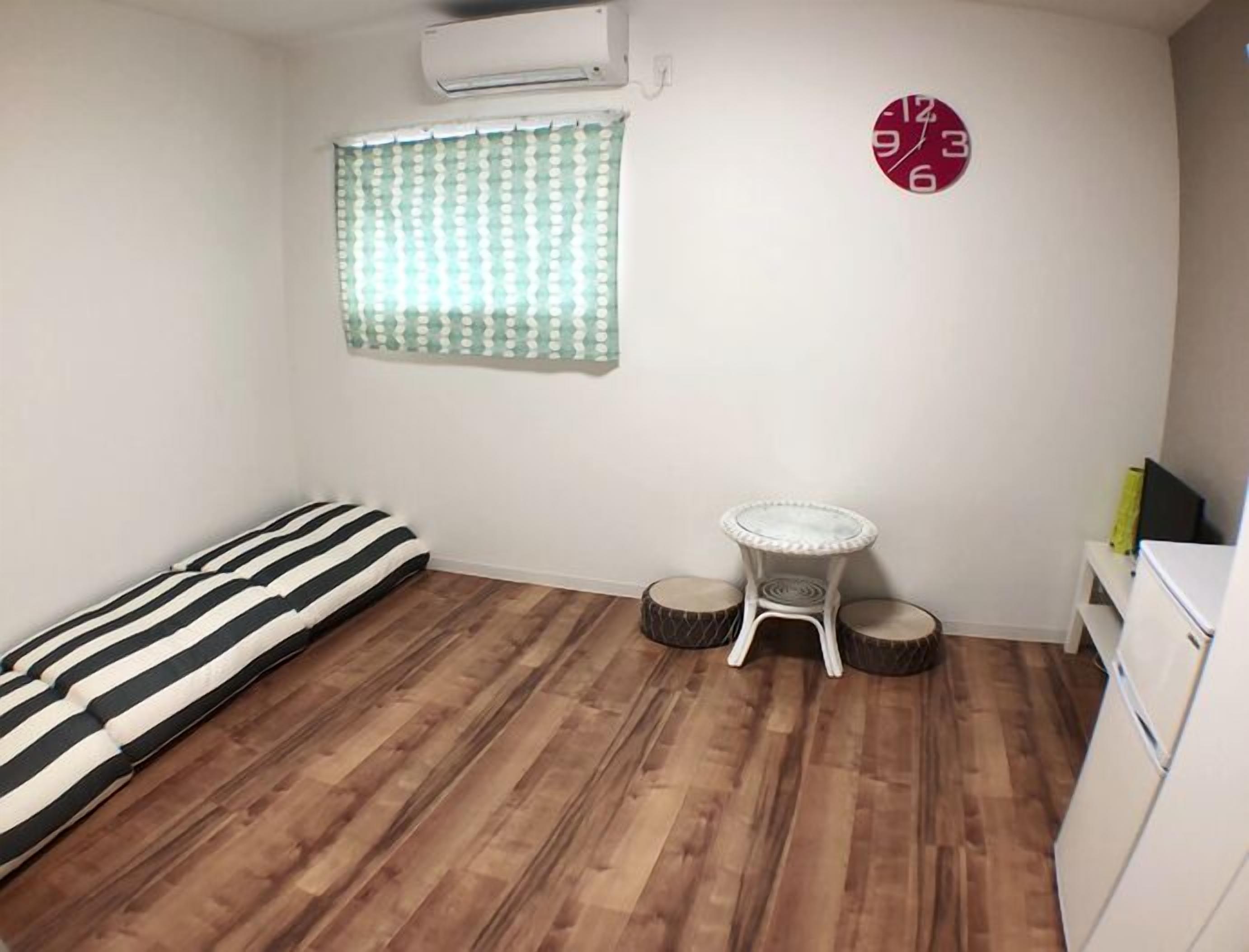 A small room with a wooden floor, a bed with a striped pillow, a small round table, and a clock on the wall.