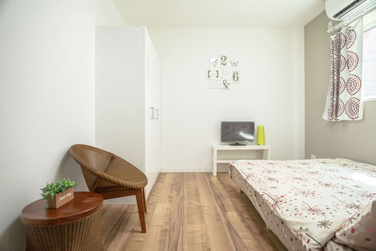Un dormitorio con cama, una silla, una mesita con una planta, un televisor sobre un mueble bajo y un armario.