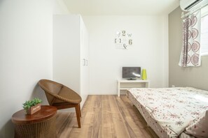 A bedroom with a bed, a chair, a small table with a plant, a TV on a stand, and a wardrobe.