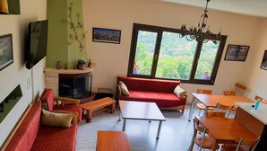 A cozy living room with a red sofa, wooden coffee table, and a fireplace. There is a dining area with a table and chairs, and a large window offering a view of greenery. - Guest House Ilektra (Voio)