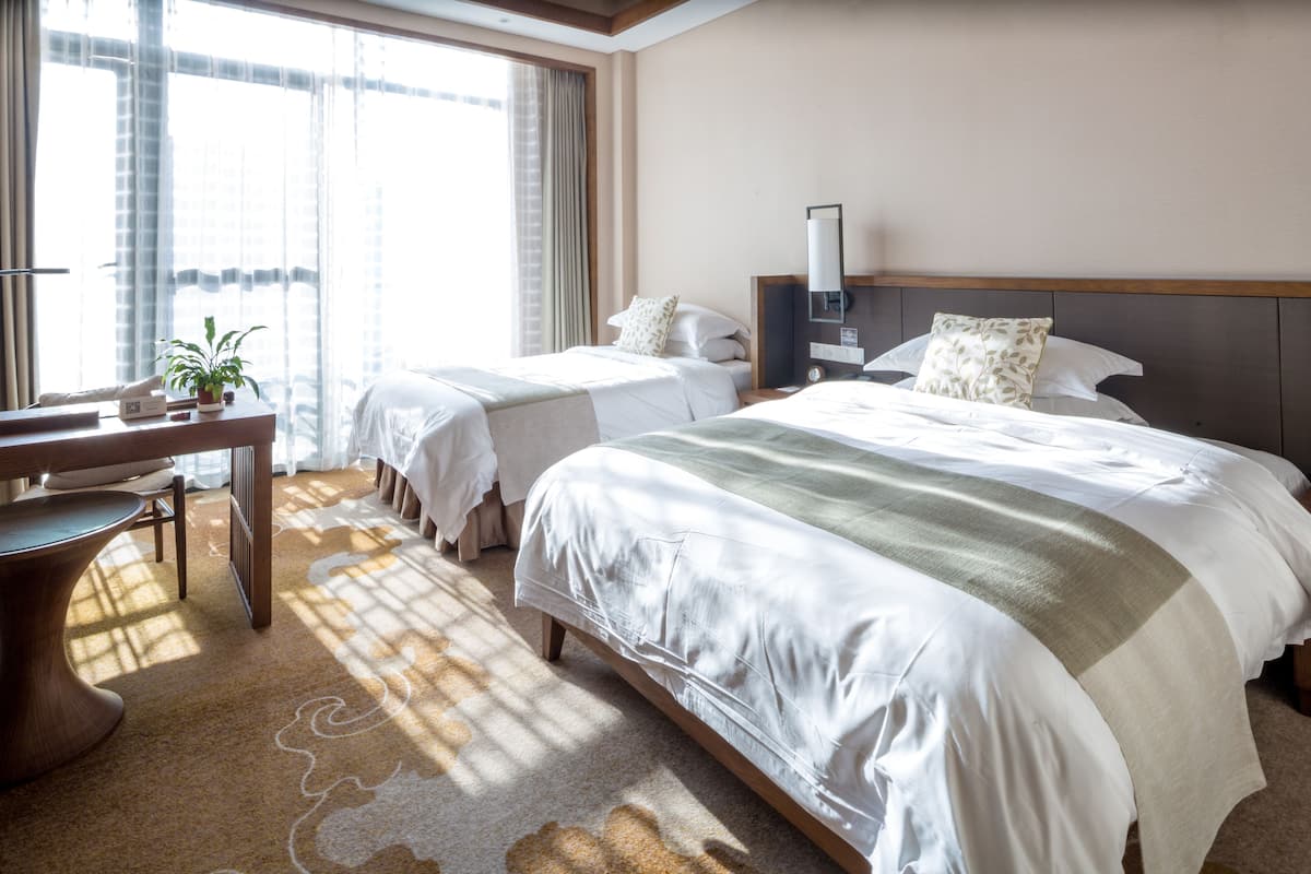 Ein Hotelzimmer mit zwei Betten, einem Schreibtisch, einem Stuhl, einer Zimmerpflanze und einem großen Fenster mit Vorhängen.