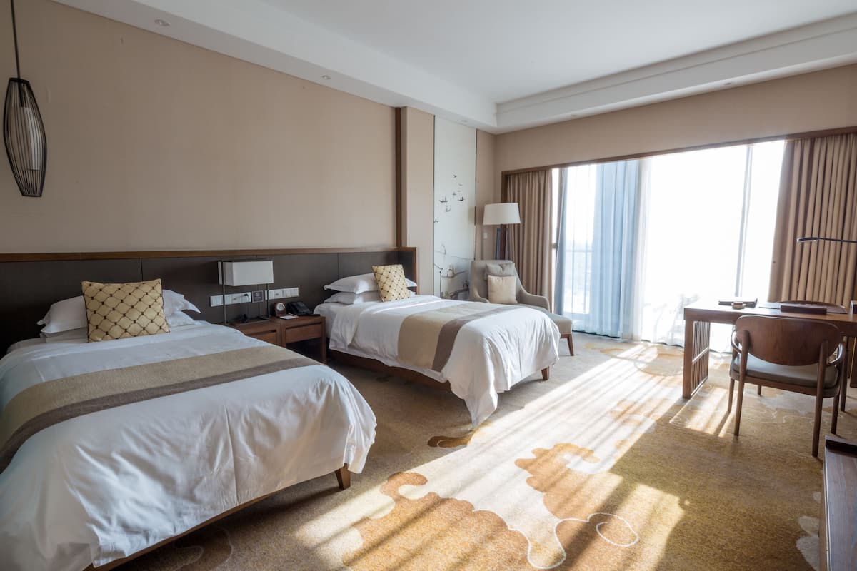 Ein Hotelzimmer mit zwei Betten, einem Schreibtisch, einem Stuhl, einer Lampe und einem großen Fenster mit Vorhängen.