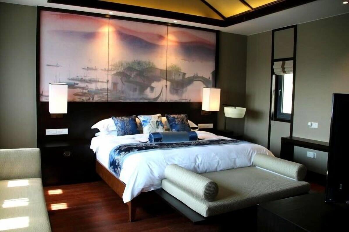Ein Hotelzimmer mit einem großen Bett, einem Schreibtisch und einem Stuhl. An der Wand hängt ein Gemälde und auf dem Schreibtisch steht eine Lampe.