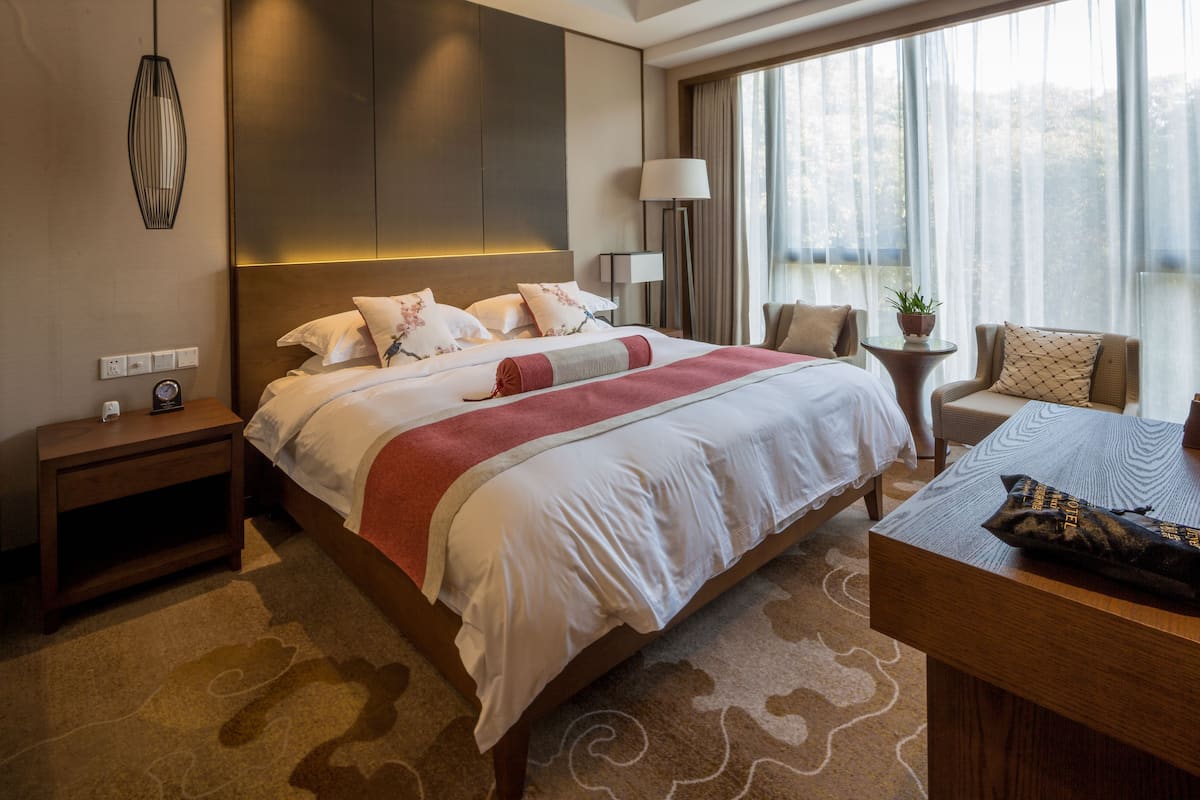 Ein Hotelzimmer mit einem großen Bett, einem Nachttisch mit Lampe, einem kleinen Beistelltisch mit Pflanze und Blick ins Freie durch große Fenster.