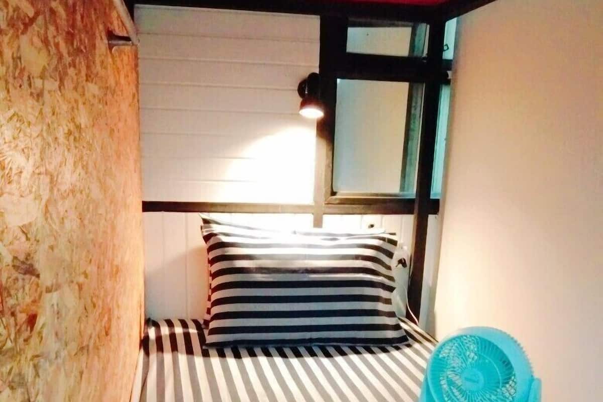 Una habitación pequeña y funcional con una cama a rayas, una ventana, un ventilador y una toalla con un estampado floral.
