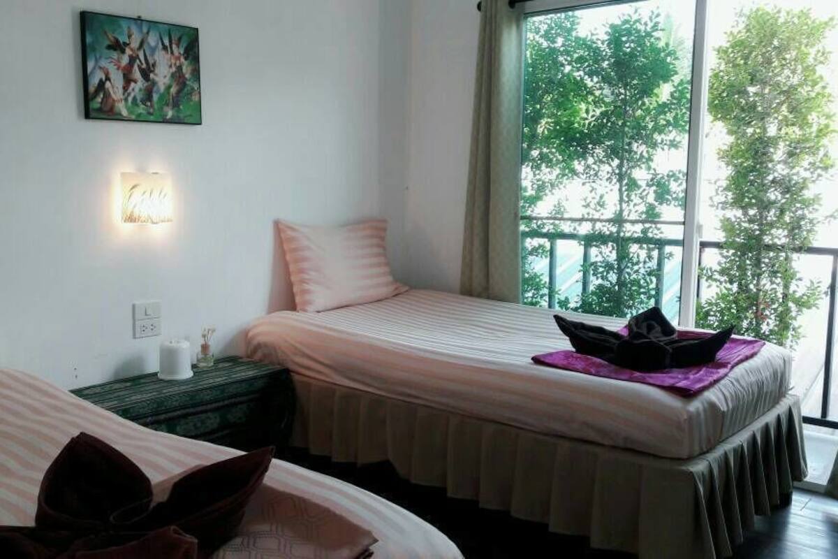 Habitación de hotel con dos camas, una mesita de noche, una lámpara de pared y un balcón con vista a árboles.