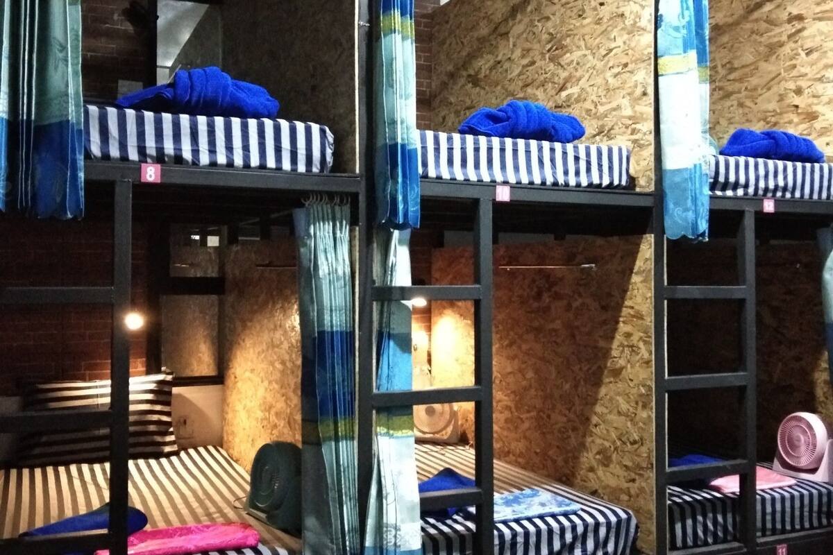 Camas literas con ropa de cama a rayas, una pared de madera y un ventilador en el suelo.
