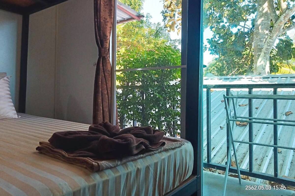Un dormitorio con cama, ventana con cortinas y vistas a un balcón y árboles.