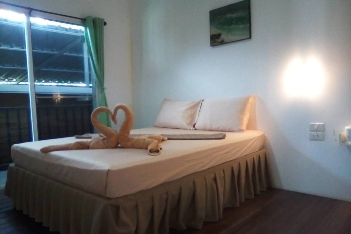Una cama bien hecha con una toalla dispuesta en forma de corazón, una mesita de noche y una ventana con vistas.
