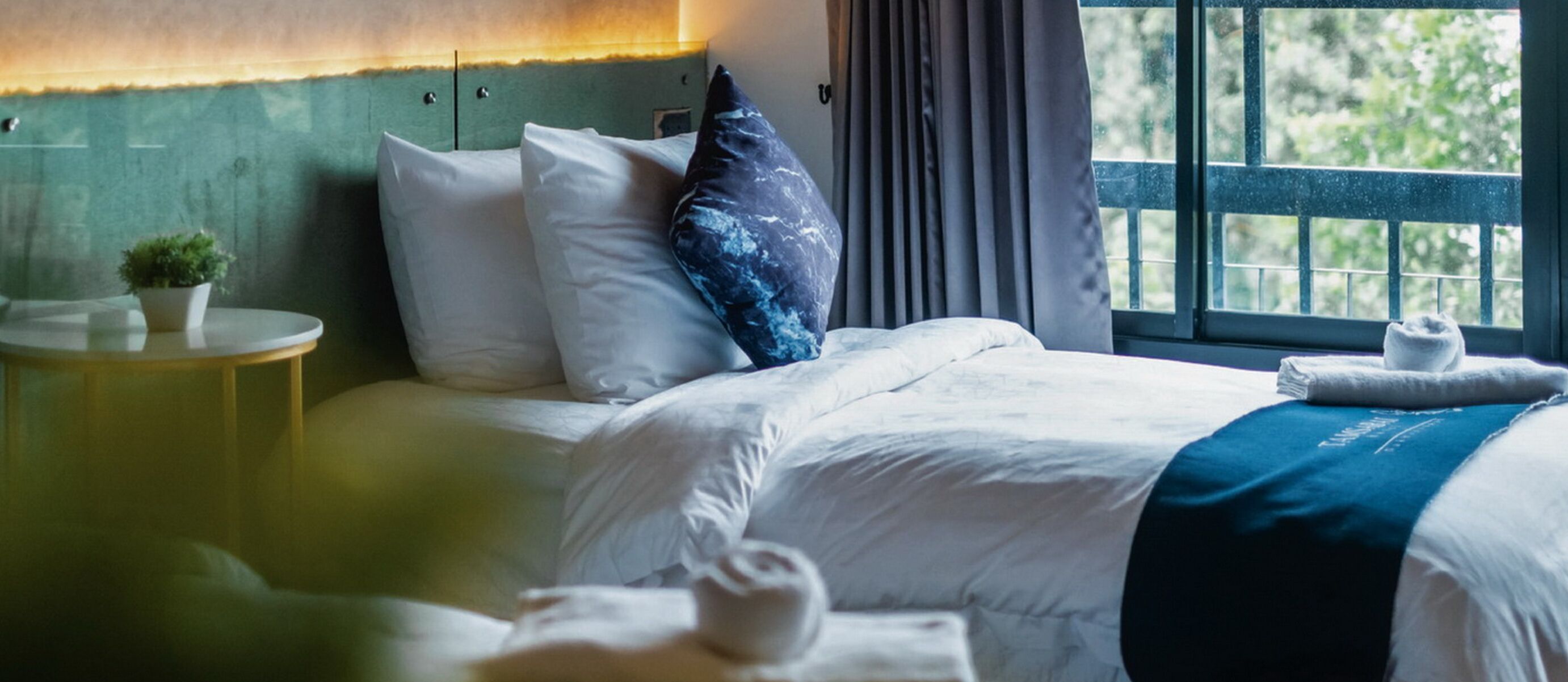 A neatly made bed with white linens and a blue throw blanket, a small round table with a potted plant, and a window with a view of greenery.