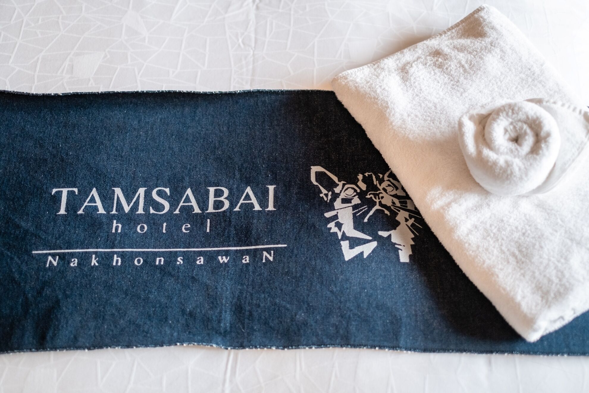 En handduk med Tamsabai Hotels logotyp och ett kattmotiv, placerad på en säng med ett vitt, strukturerat sängöverkast.