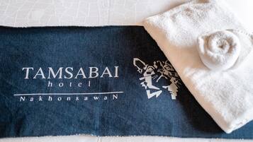 En handduk med Tamsabai Hotels logotyp och ett kattmotiv, placerad på en säng med ett vitt, strukturerat sängöverkast.