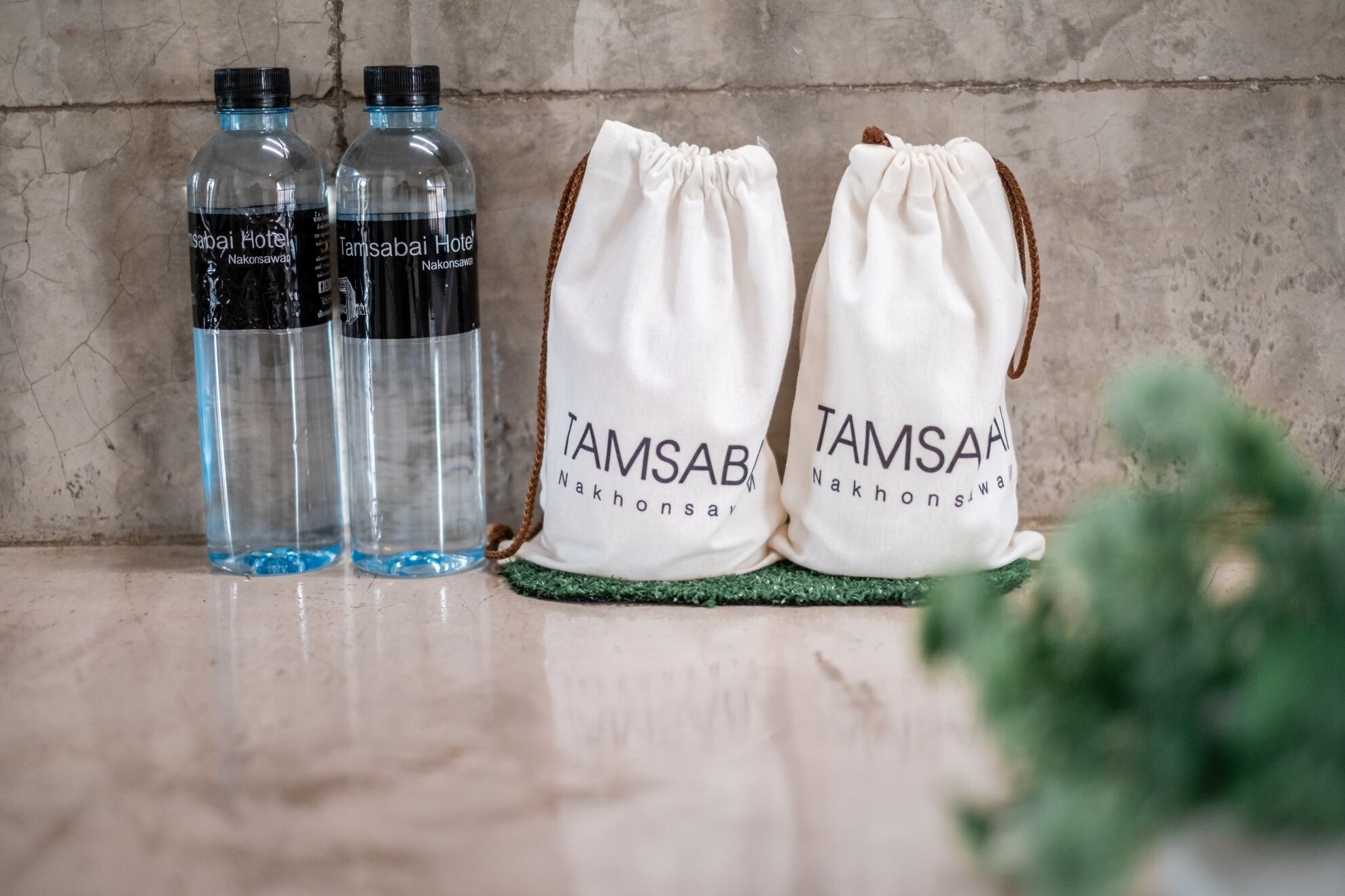 Trois bouteilles d’eau transparentes et deux sacs blancs à cordon de serrage, portant l’inscription « Tamsabai Hotel Nakhonsawai », sont posés sur une surface texturée.