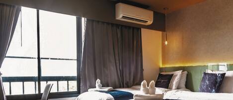 A hotel room with two beds, a chair, a window with curtains, an air conditioning unit, and a wall-mounted light fixture.