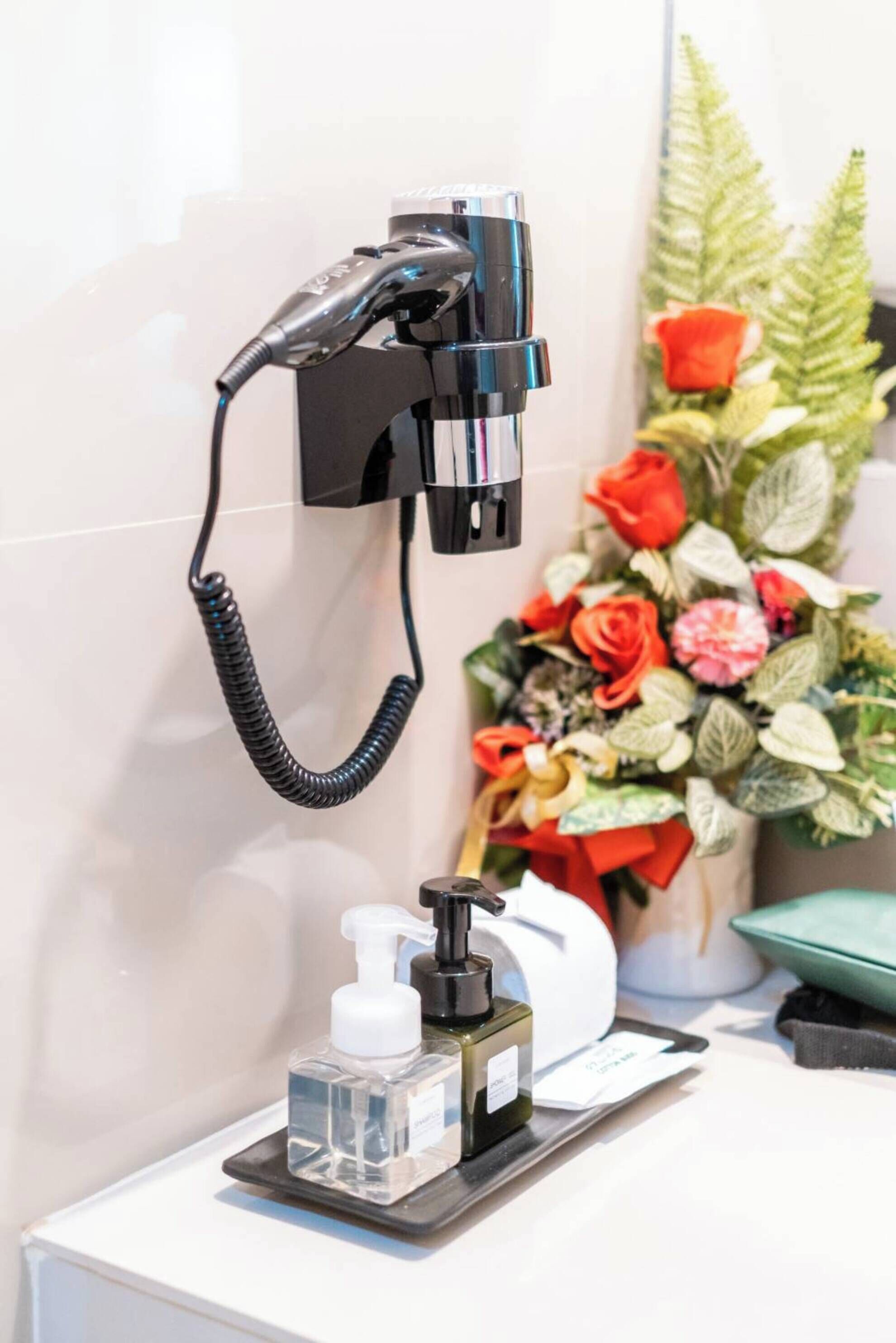 A bathroom with a hairdryer mounted on the wall, a soap dispenser, and a floral arrangement on the counter.