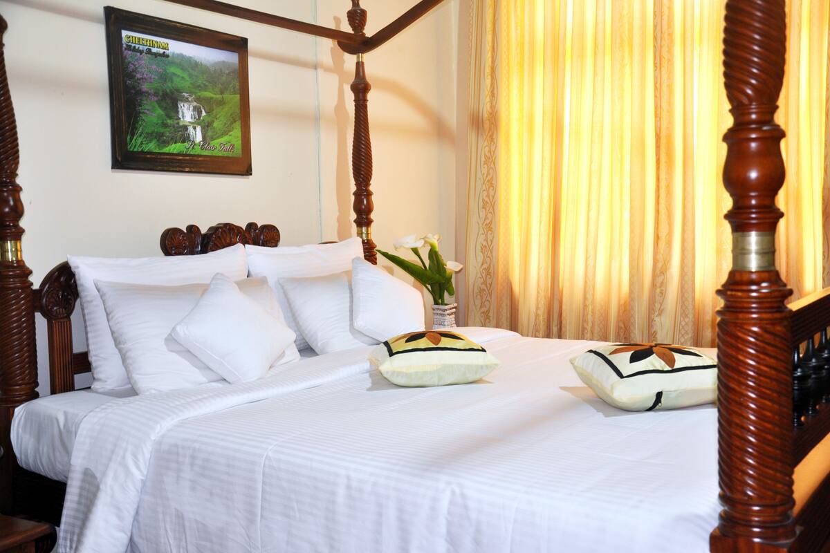 A four-poster bed with white linens and decorative pillows, a framed picture on the wall, and a vase with flowers on the nightstand.