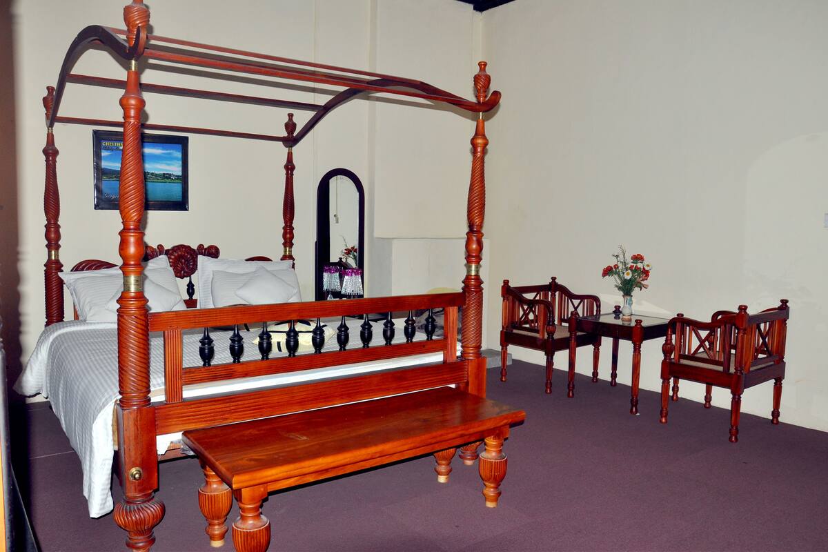 A four-poster bed with a wooden bench in front, a dining table with chairs, and a mirror on the wall.
