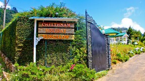 Un letrero para Cheltnam Holiday Bungalow con un diseño rústico, rodeado de exuberante vegetación y un cielo despejado.