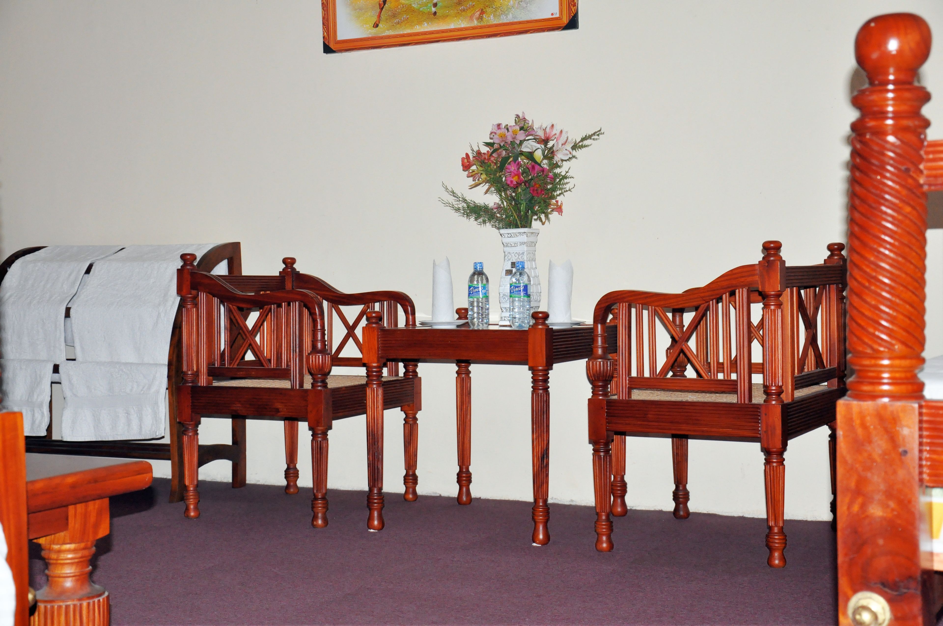 A room with a wooden dining table, two chairs, a vase with flowers, and a bed with a white blanket.