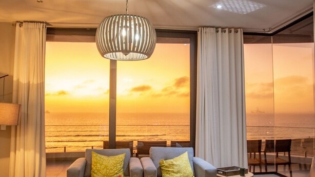 Ein modernes Wohnzimmer mit großem Fenster und Meerblick bei Sonnenuntergang, ausgestattet mit einer Couch, einem Tisch und einer Pendelleuchte.