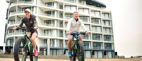 Two people riding bicycles in front of a modern building with multiple floors and balconies.