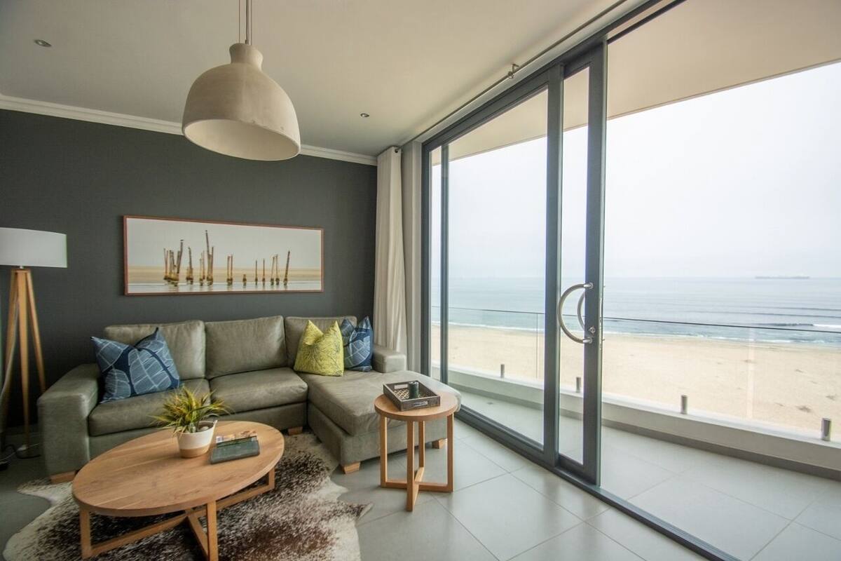 A modern living room with a large window overlooking a beach, a sofa with cushions, a coffee table, and a pendant light.