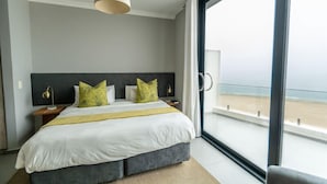 Ein modernes Schlafzimmer mit einem großen Bett, einem Nachttisch, einer Lampe und Blick auf den Strand durch ein großes Fenster.