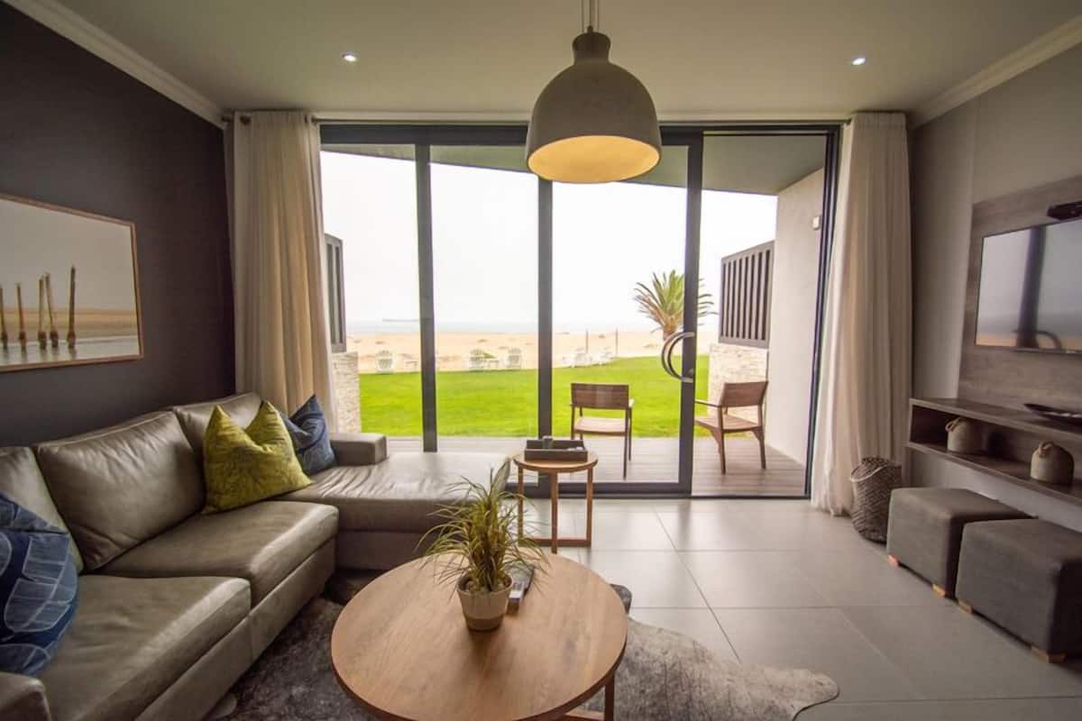 A modern living room with a sofa, coffee table, and TV. It features large windows with a view of a green lawn and a distant beach.