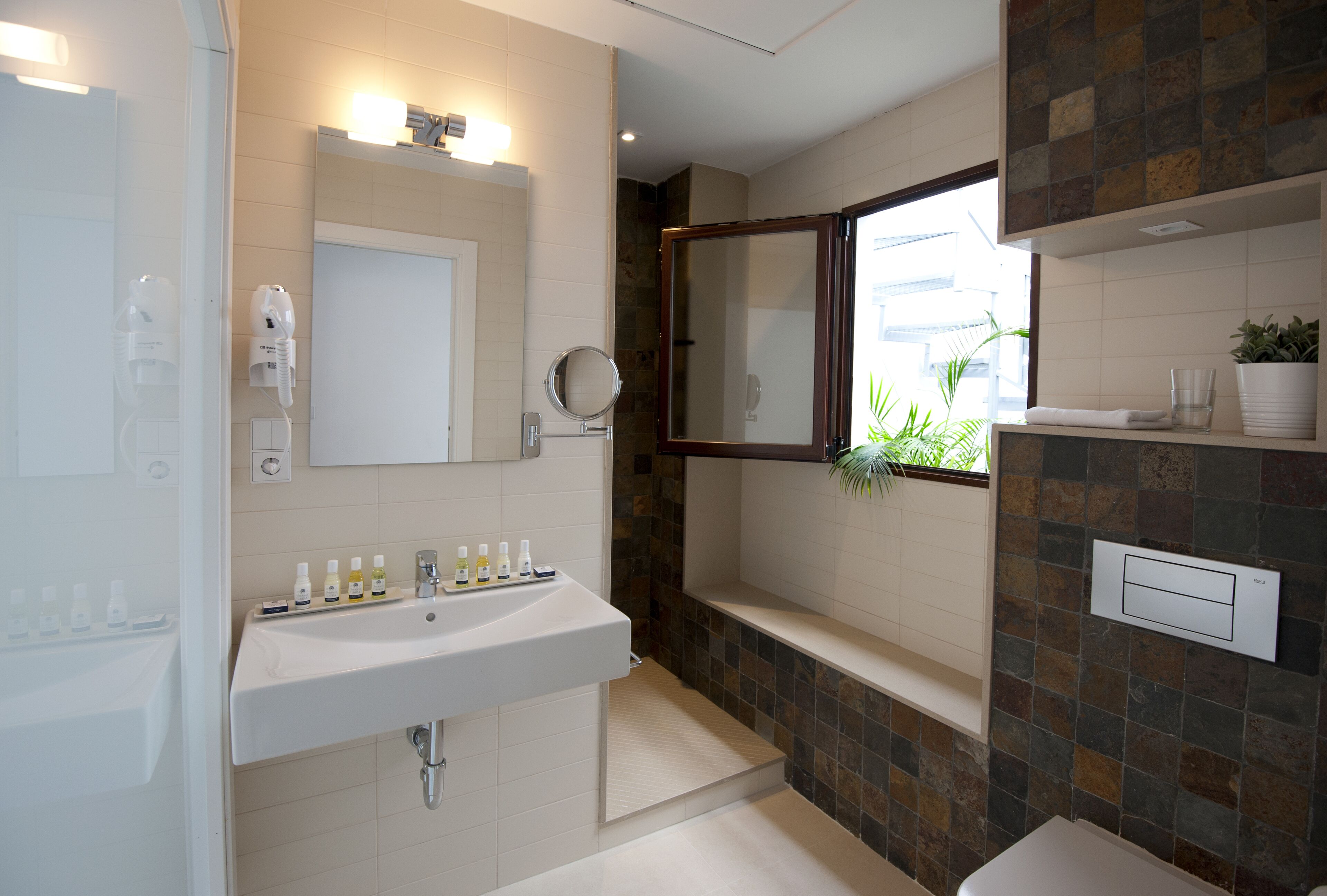 A modern bathroom with a white sink, a mirror, and a shower. There are toiletries on the sink and a potted plant on the counter.