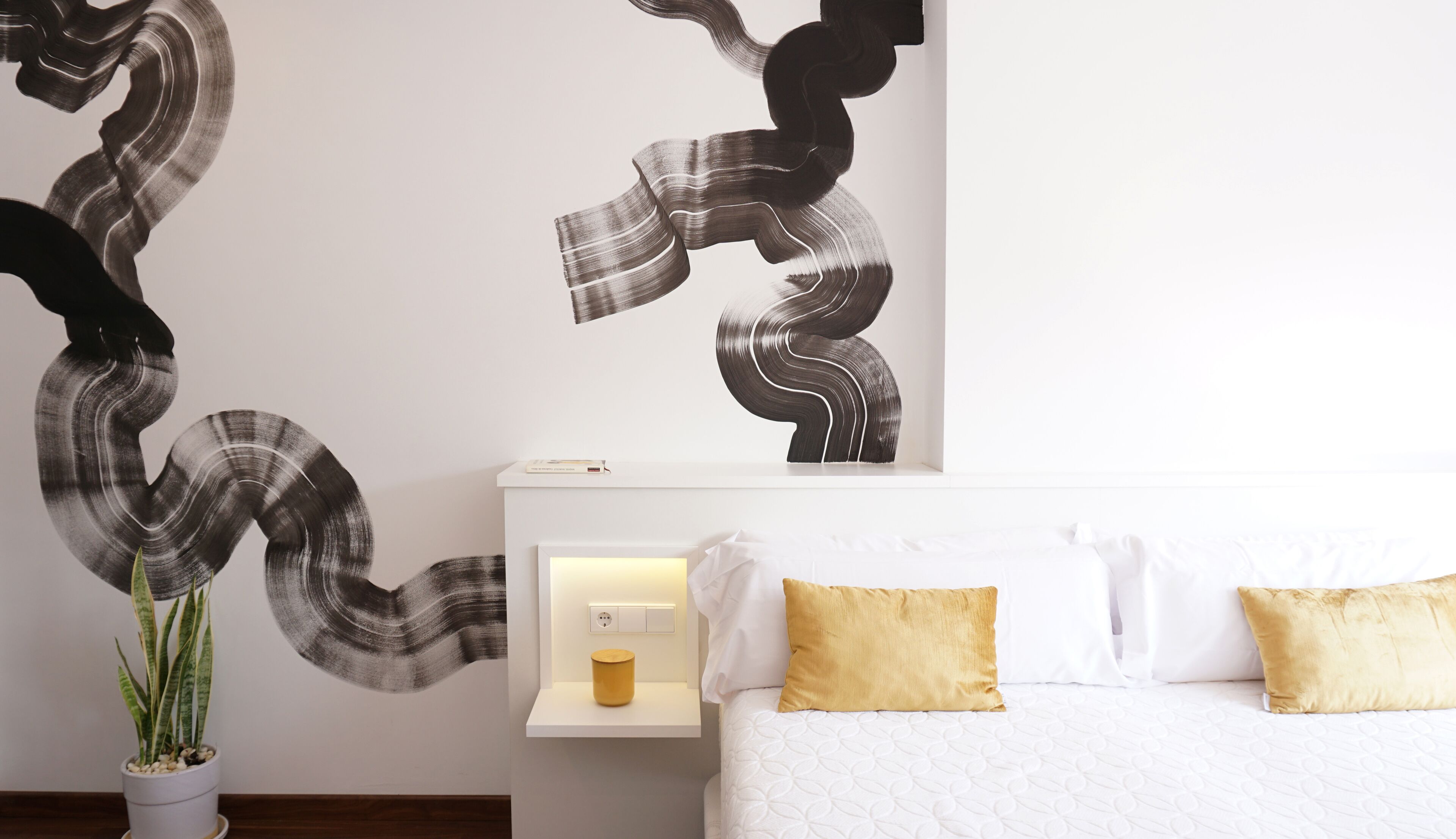 A modern hotel room with a large abstract wall art, a bedside table with a lamp, and a potted plant.