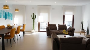 Ein geräumiges Wohnzimmer mit einem braunen Sofa, orangefarbenen Sesseln und einem Esstisch mit Obstschale. Auf dem Tisch stehen Kakteen in Töpfen und eine Vase mit Blumen.